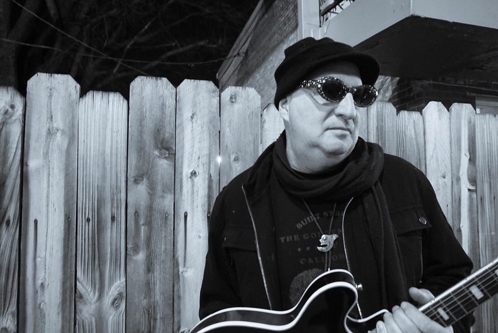 black and white photo shows keith scott in front of a fence holding a guitar, looking moody